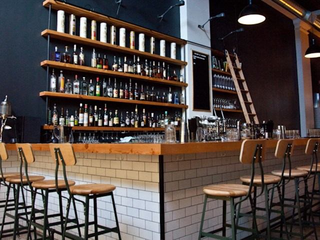 Rustic wood bar with stools, shelves of bottles, and warm hanging lights.