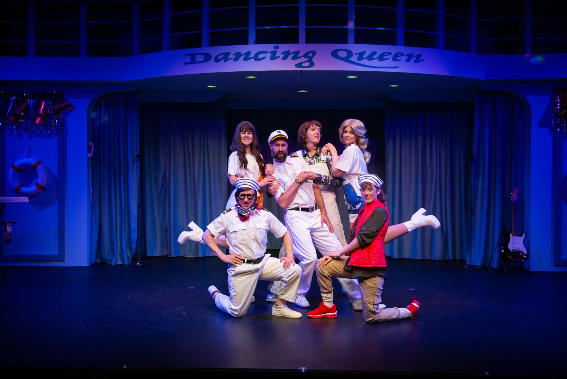 Stage performers in sailor costumes pose under Dancing Queen sign on cruise ship set.