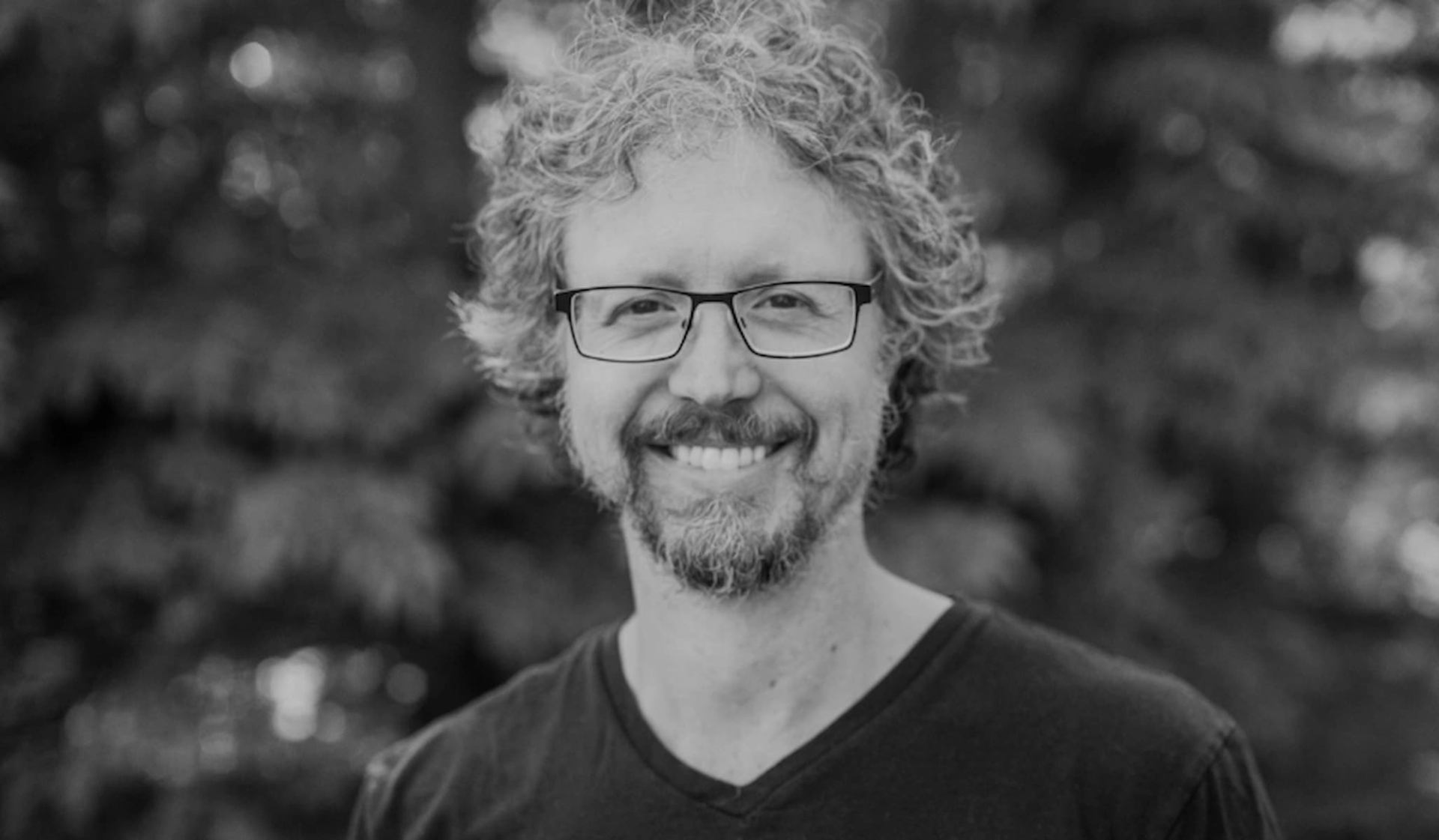 Person with curly hair and glasses stands outdoors in front of blurred trees.
