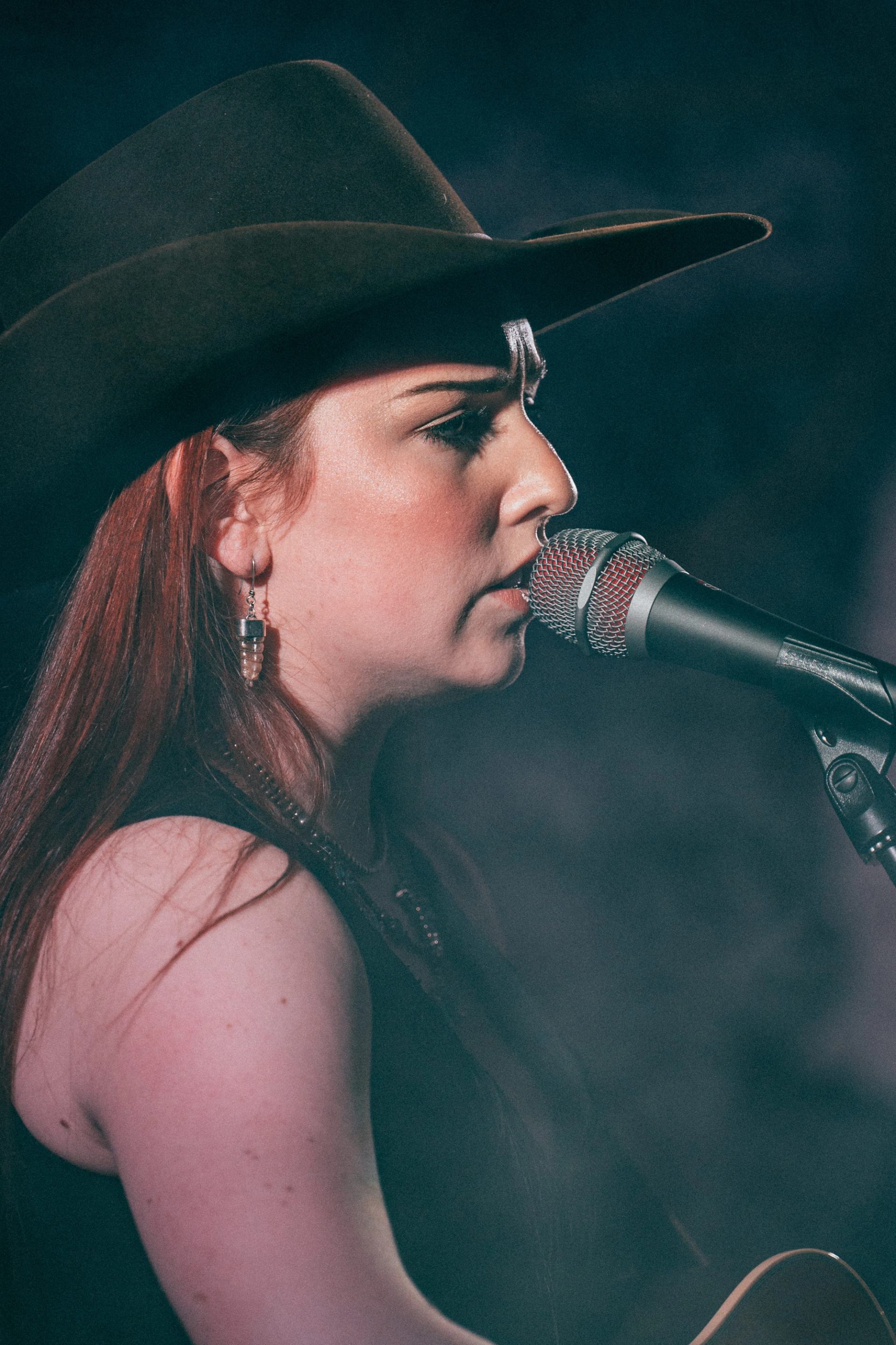 Performer singing into a microphone while playing guitar, wearing a wide-brimmed hat