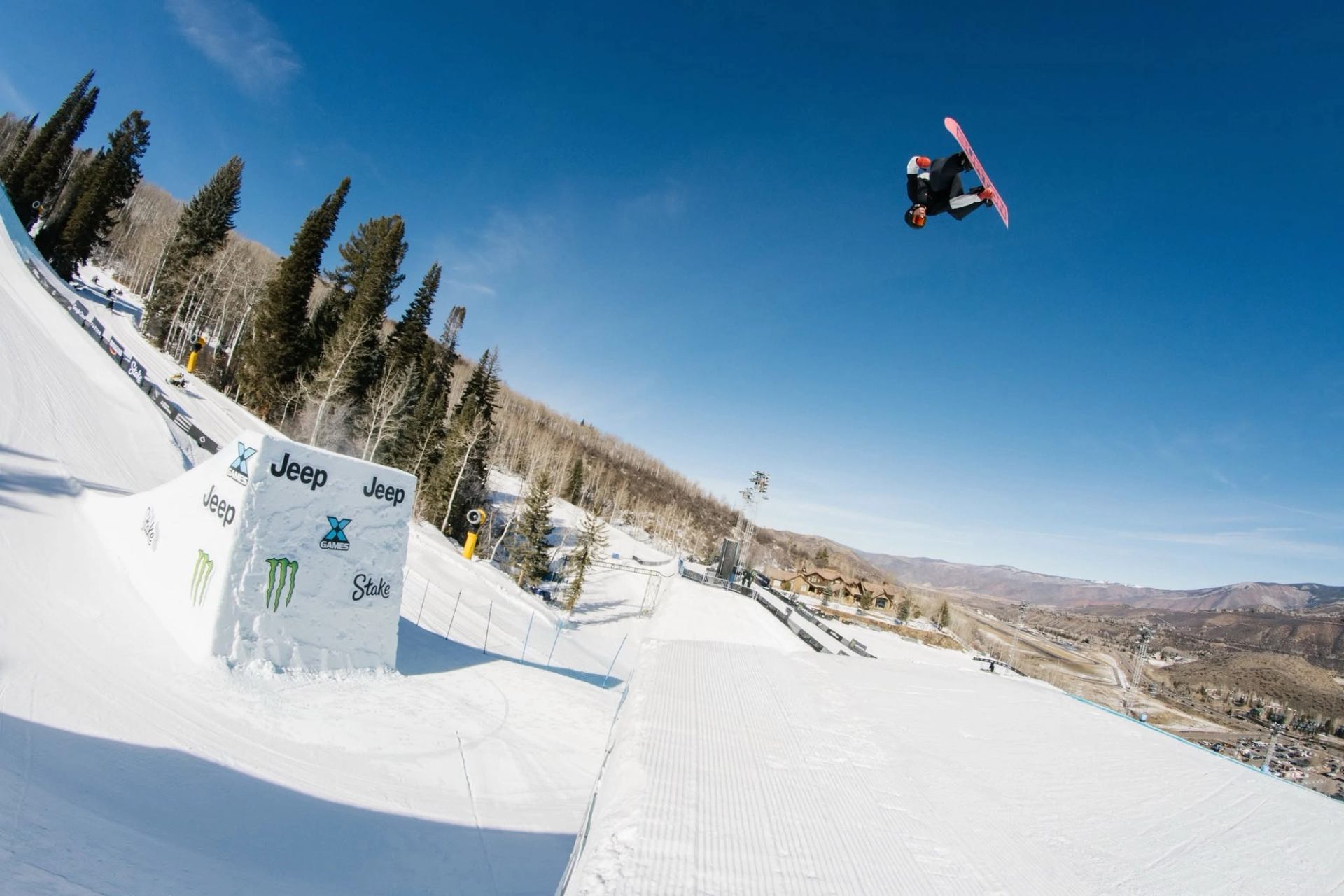 Snowboarder performs an aerial trick above a snowy course lined with trees.