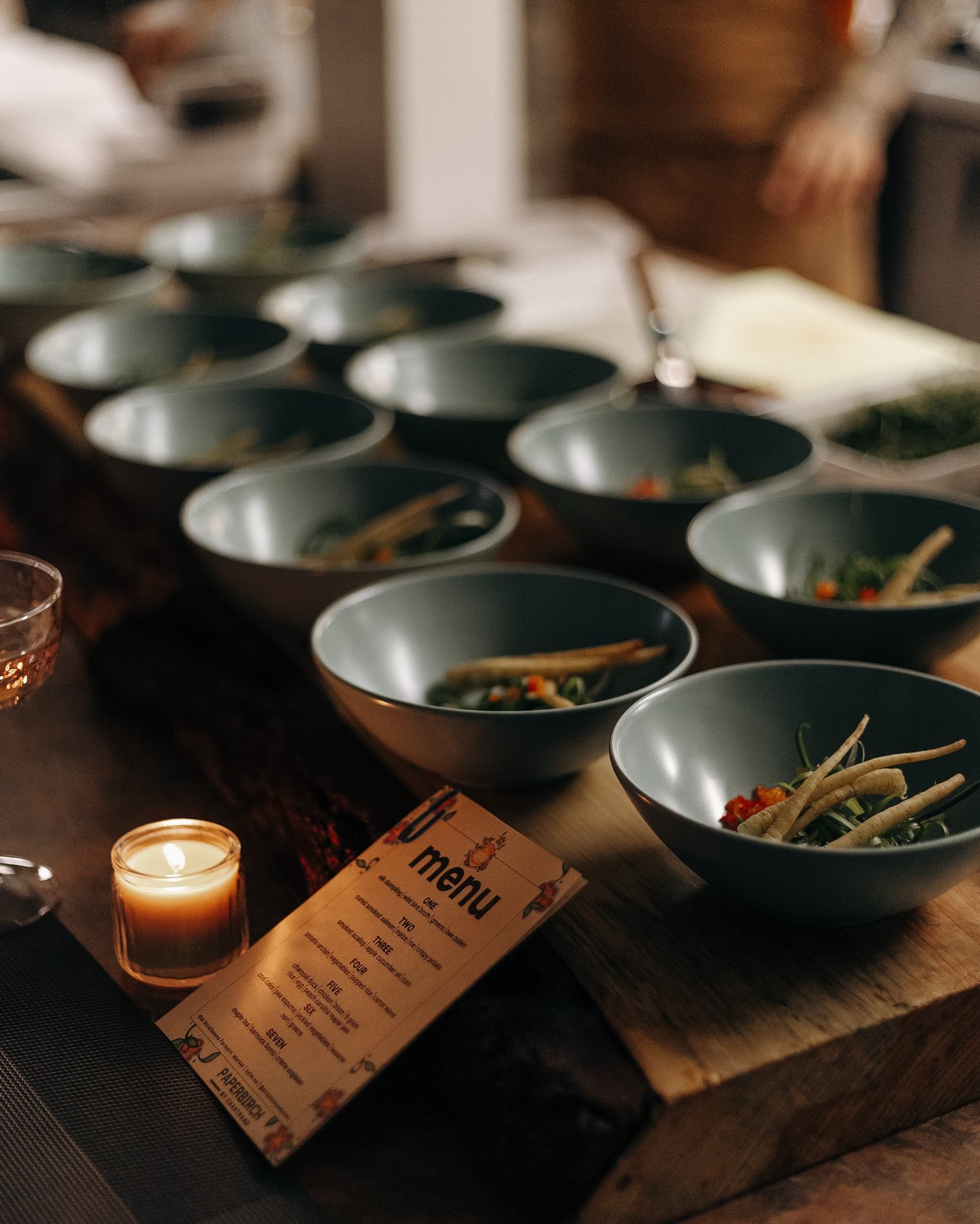 Row of prepared salad bowls on wooden counter beside candle and printed menu.