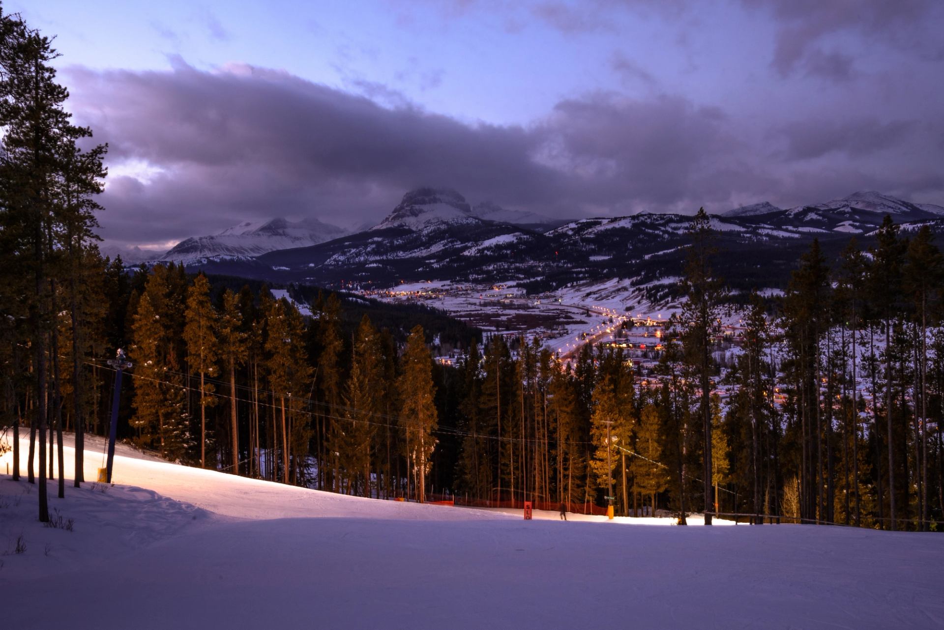 Snow-covered ski run at dusk overlooking a lit mountain valley with trees and surrounding peaks.