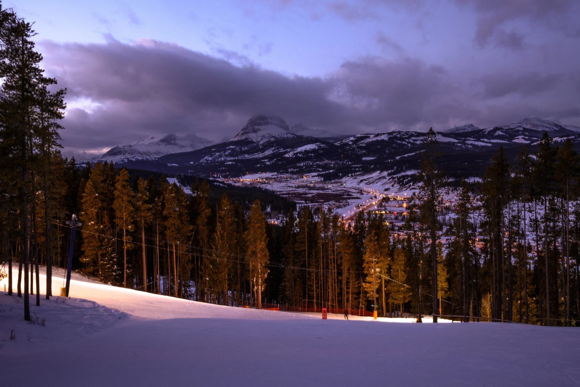 Snow-covered ski run at dusk with mountain peaks and valley lights at Pass Powderkeg.
