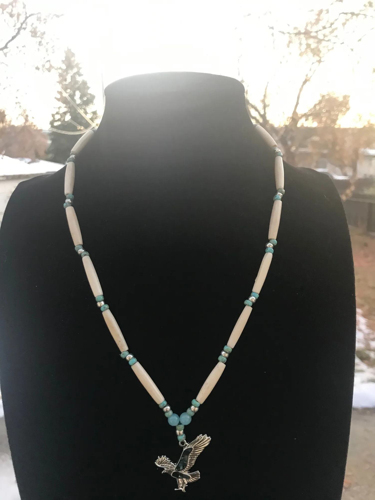 Beaded necklace with silver bird pendant displayed on black bust outdoors.