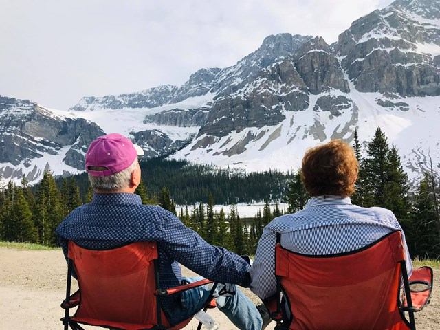 People sitting in red camp chairs facing snow-covered mountains and evergreen forest.