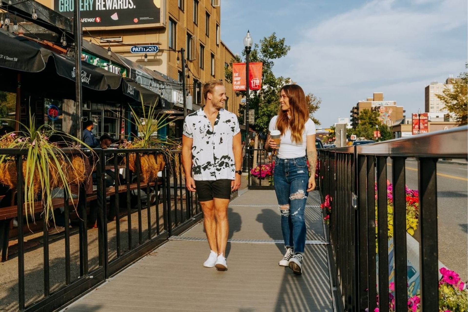 Two people walk on a raised sidewalk beside patios, planters and city street traffic.