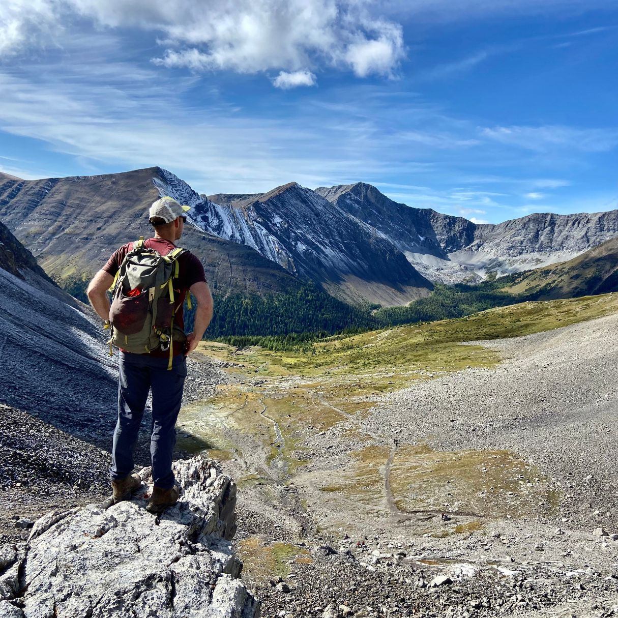 Hiker with backpack stands on rocky ridge overlooking mountain valley and peaks.