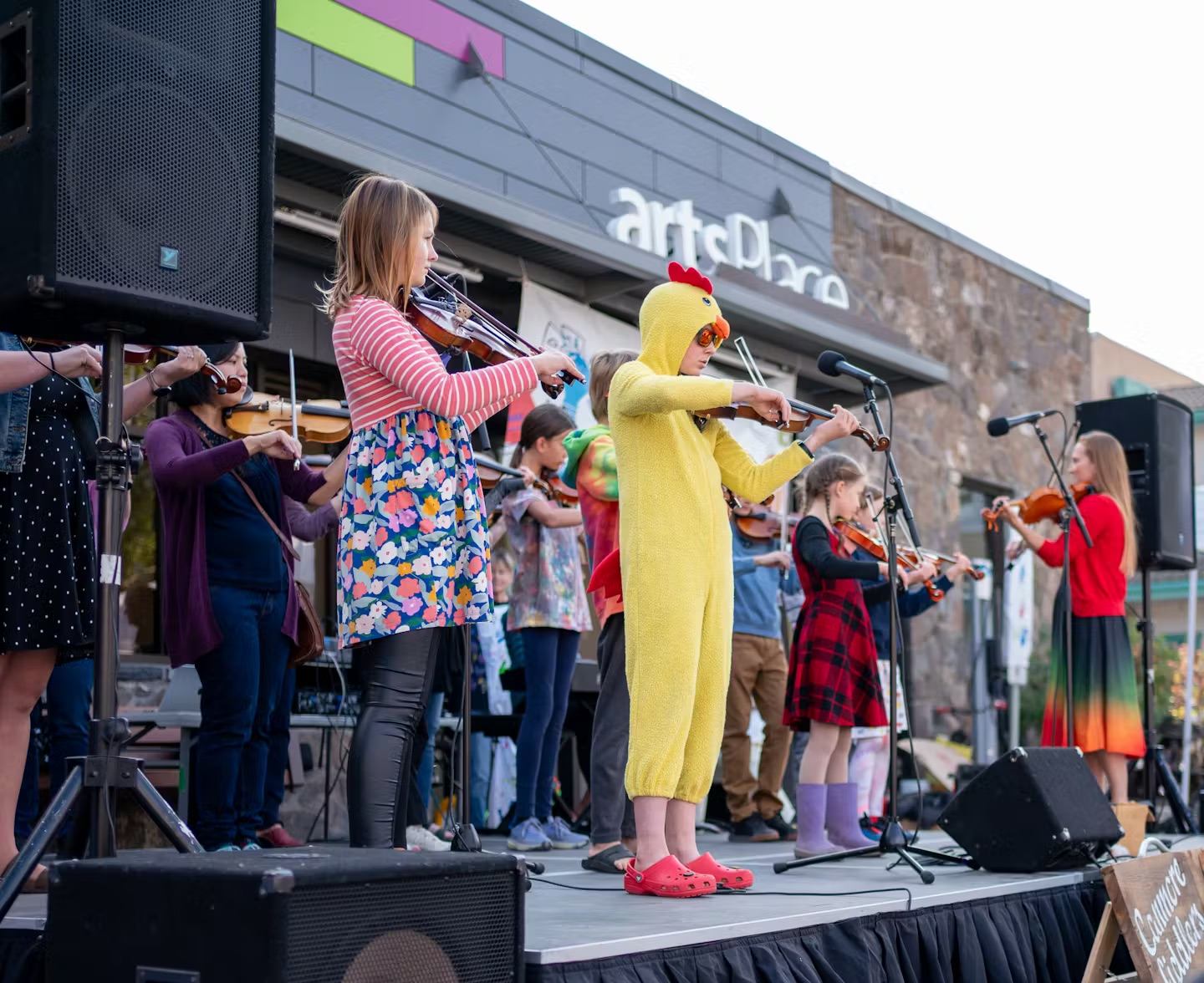 Children play violins on an outdoor stage, one dressed in a yellow chicken costume for fun.
