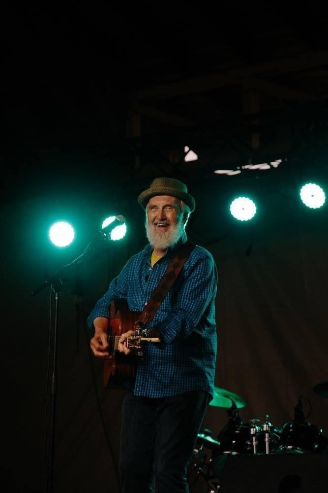 Musician in hat plays acoustic guitar on stage under green concert lights.