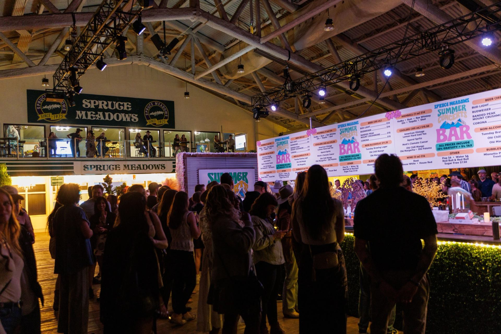 Indoor crowd at Spruce Meadows bar, with large illuminated drink menu overhead.