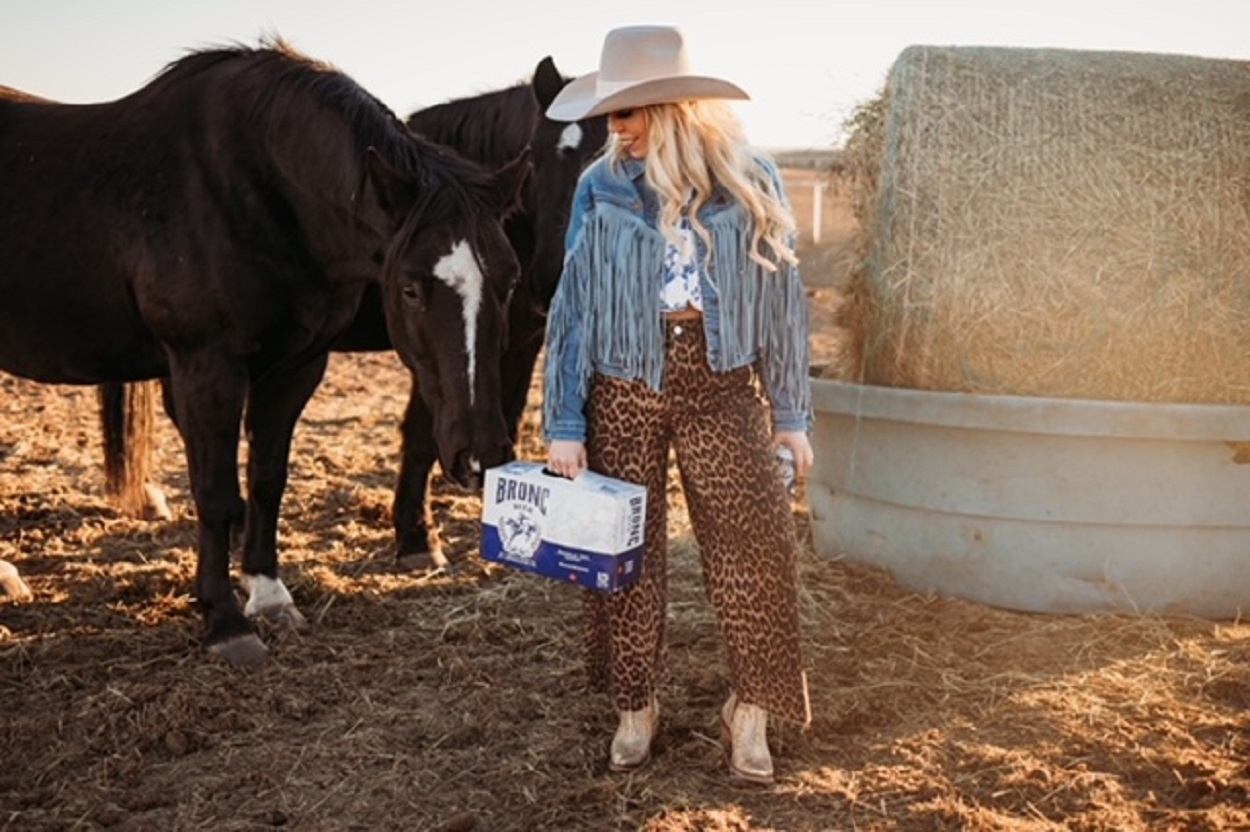 A cowgirl holding a case of Bronc Beer with two horses behind her