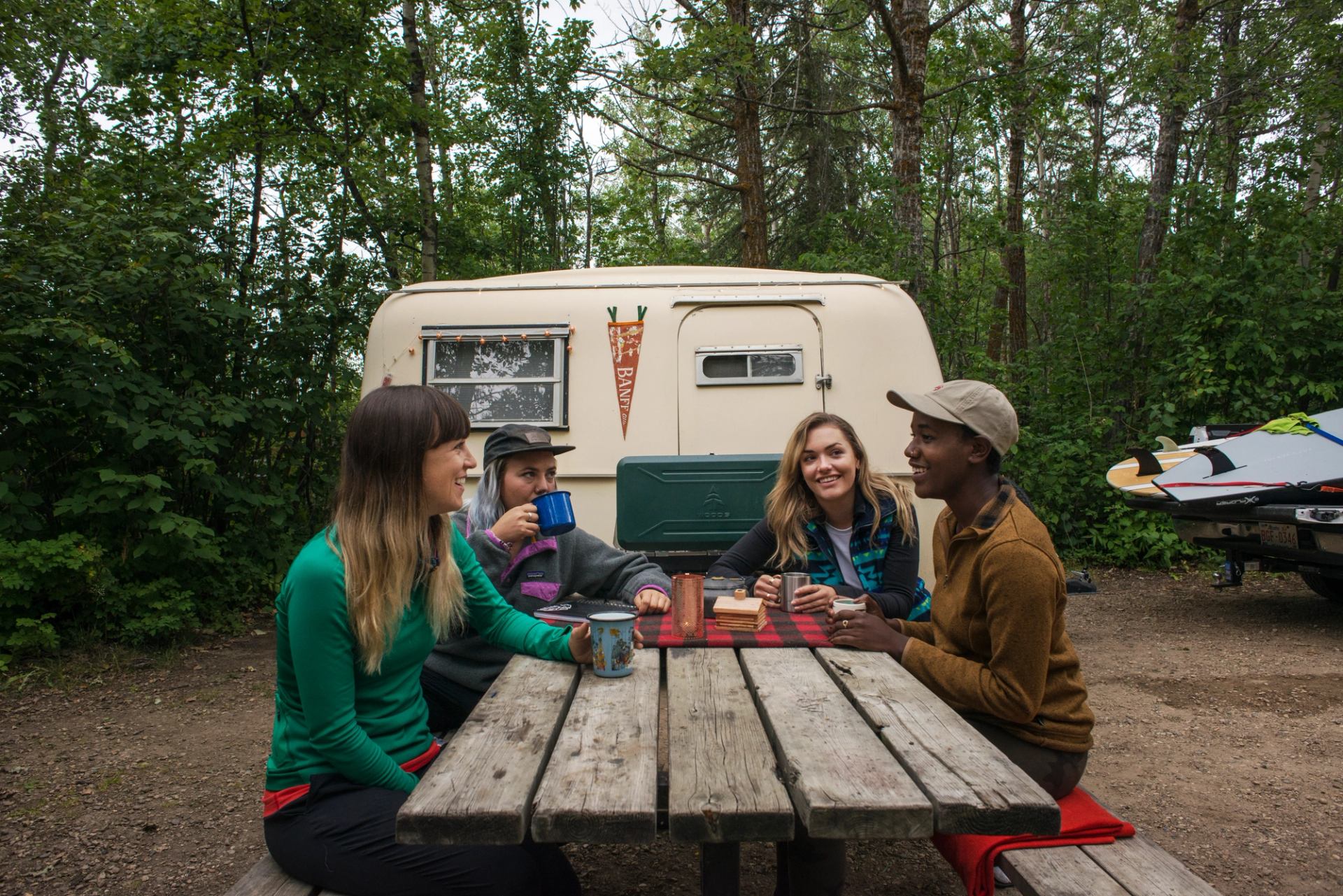 Group sits at picnic table by small trailer, drinking from mugs in forest campground.