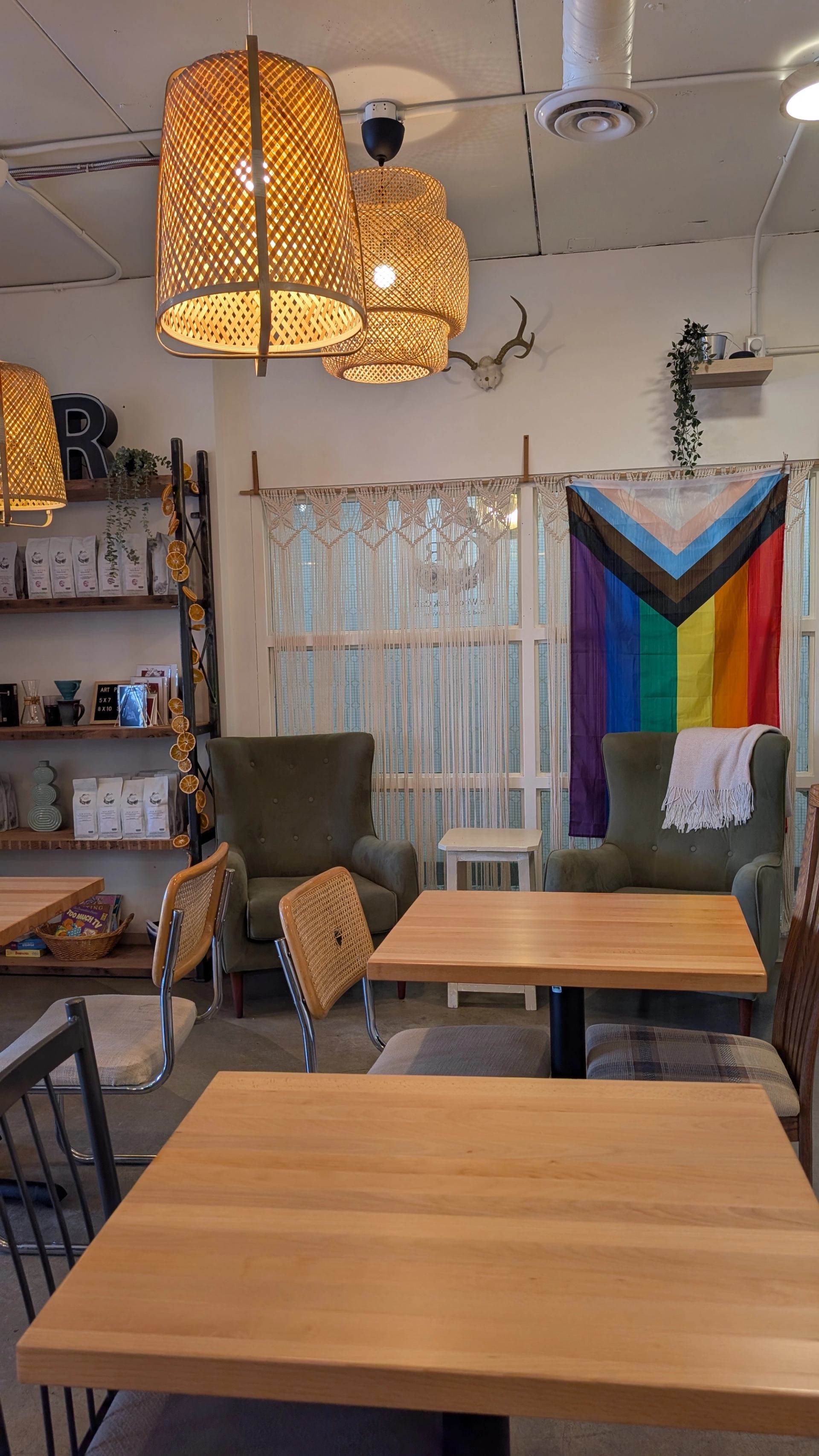 Cafe seating area with wooden tables, armchairs, woven lights and Progress Pride flag.