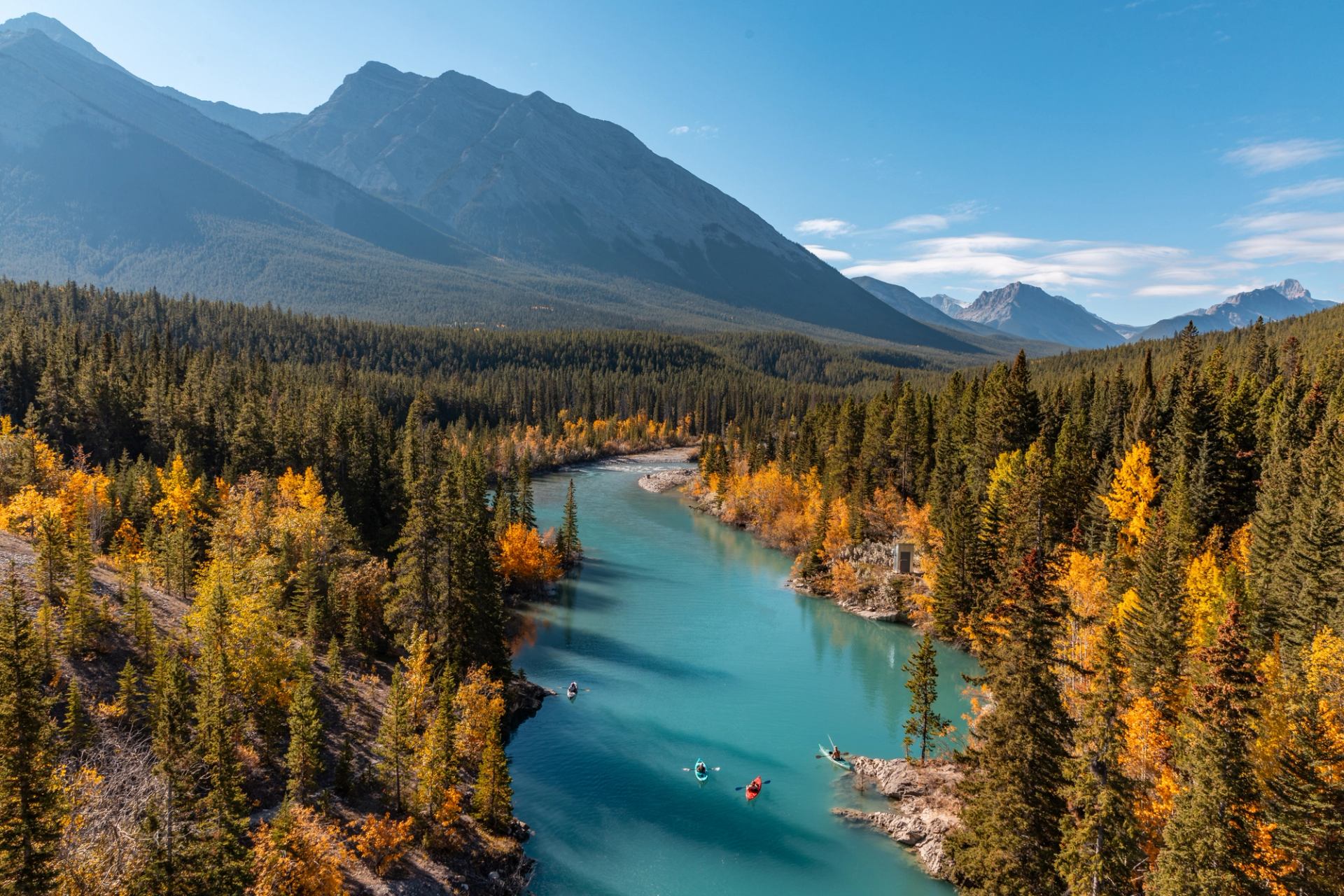 Turquoise river winding through autumn forest with mountains under a clear blue sky.
