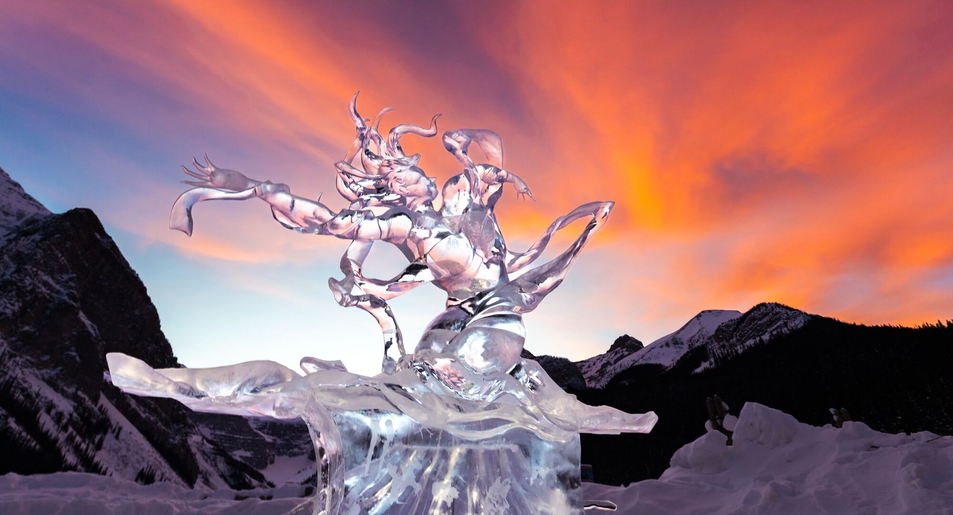 Intricate ice sculpture set against a vivid sunset sky and snow-covered mountains.
