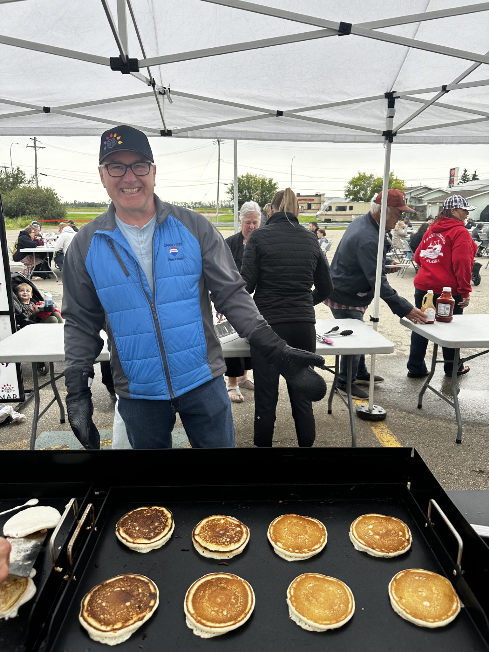 Man in blue vest cooks pancakes on outdoor griddle under canopy at community event.