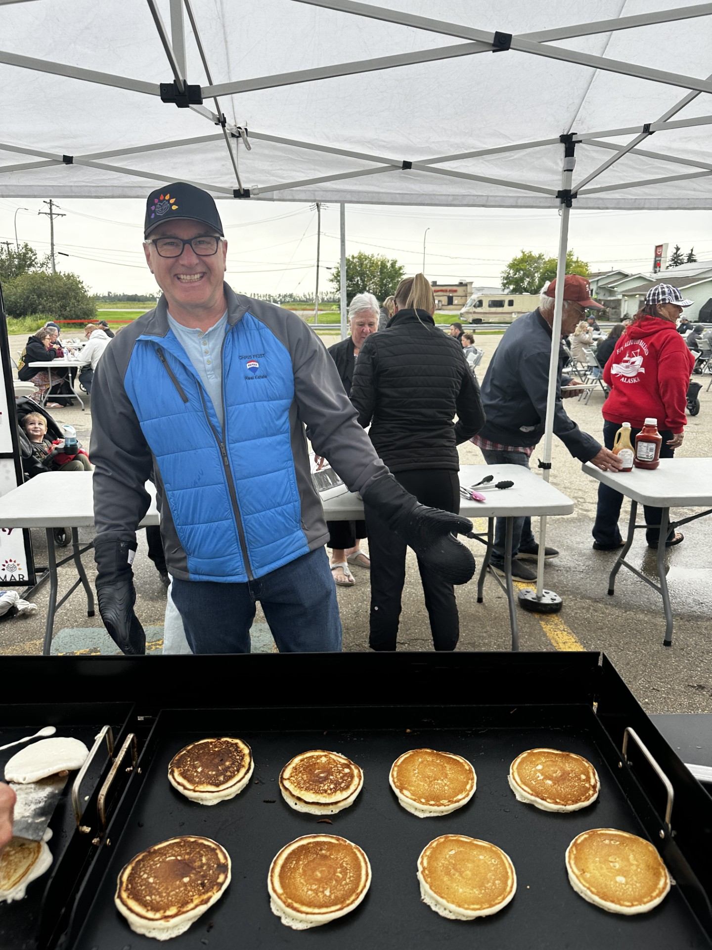 Man in blue vest cooks pancakes on outdoor griddle under canopy at community event.