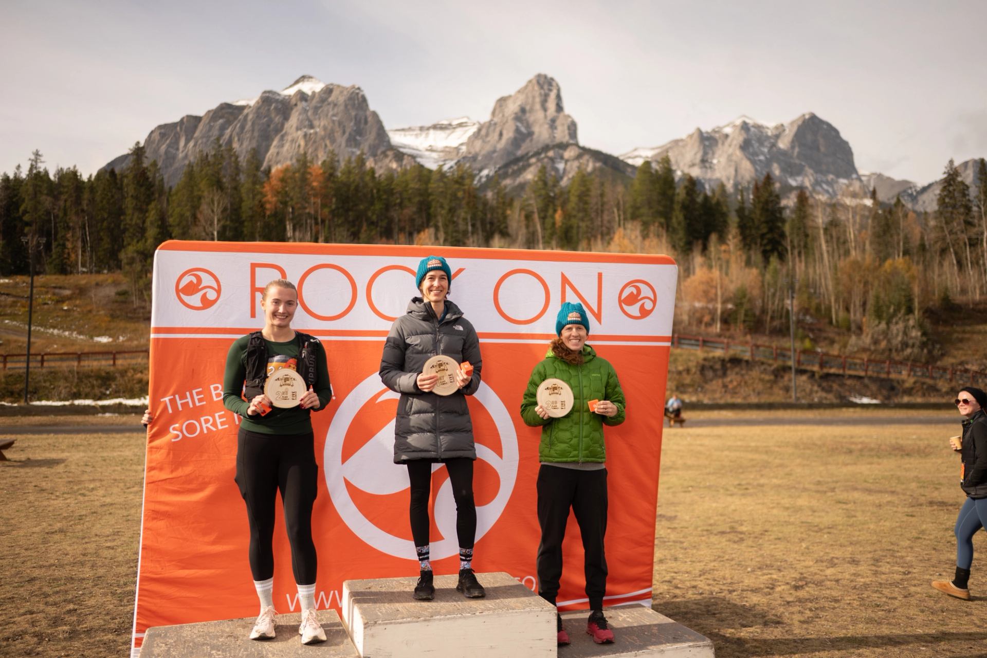 Three race winners stand on a podium holding wooden awards with mountains in the background.