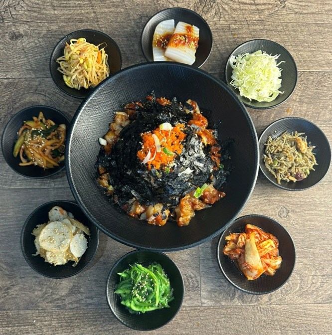A large Korean rice bowl topped with shredded seaweed and orange roe, surrounded by eight small bowls of banchan on a wooden table.