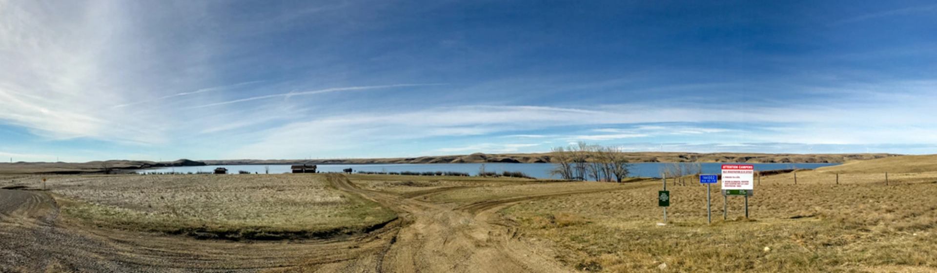 Gravel road leading to calm lake, dry grassland, and several posted information signs.