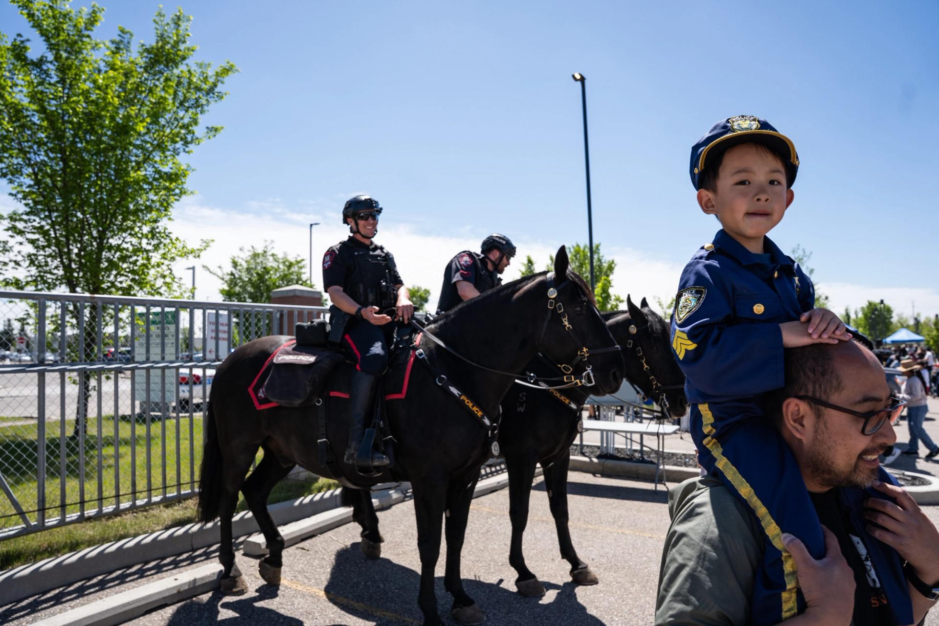 Child in police costume rides on shoulders beside mounted police officers.