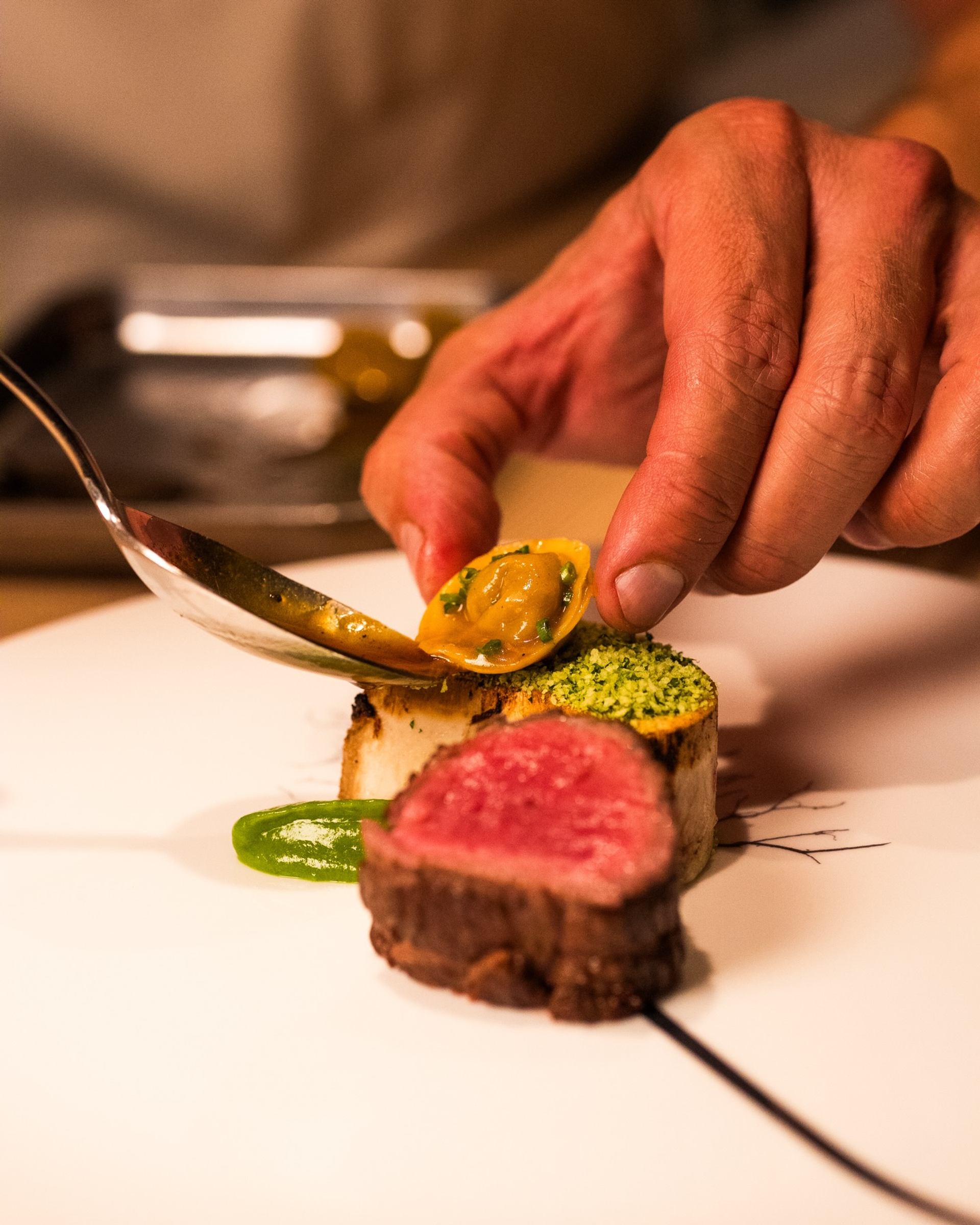 Chef’s hand plating seared beef medallion with vegetables and sauce on white dish.