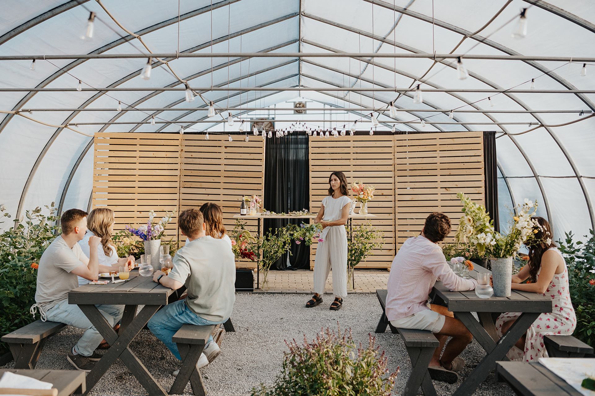 People seated at tables in a greenhouse while someone leads a workshop with flowers displayed.