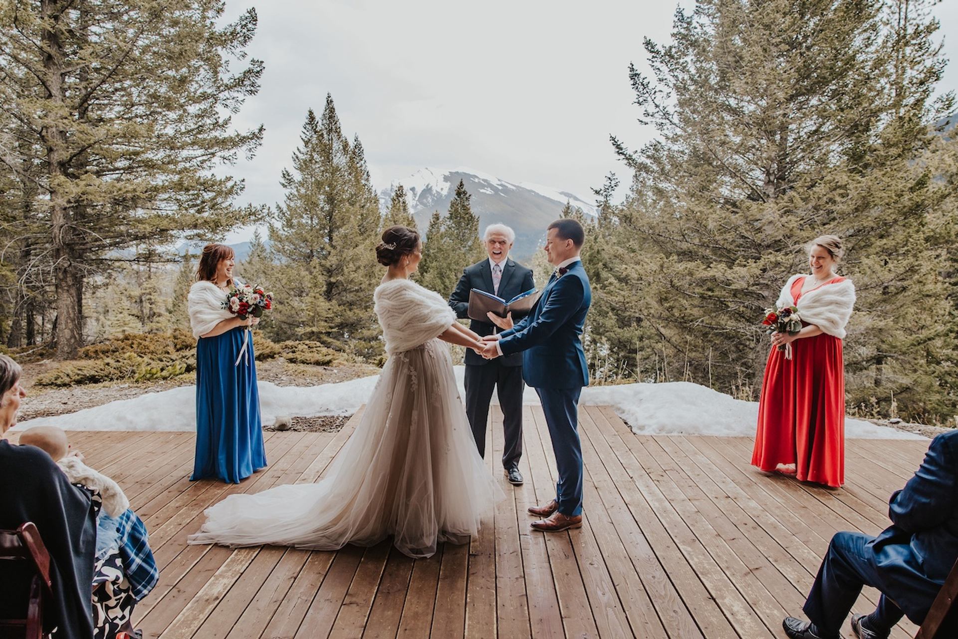 Couple standing on a wooden deck with snow, trees, and mountains behind