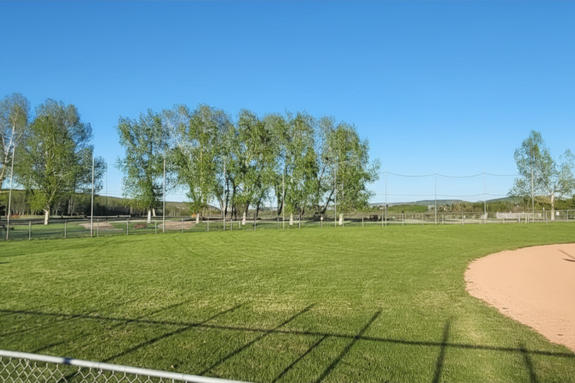 Diamond Valley Municipal Campground highlights a spacious green field bordered by trees and fencing, with a nearby baseball diamond visible to one side. The open area offers wide views under a bright blue sky, perfect for recreation and relaxation.
