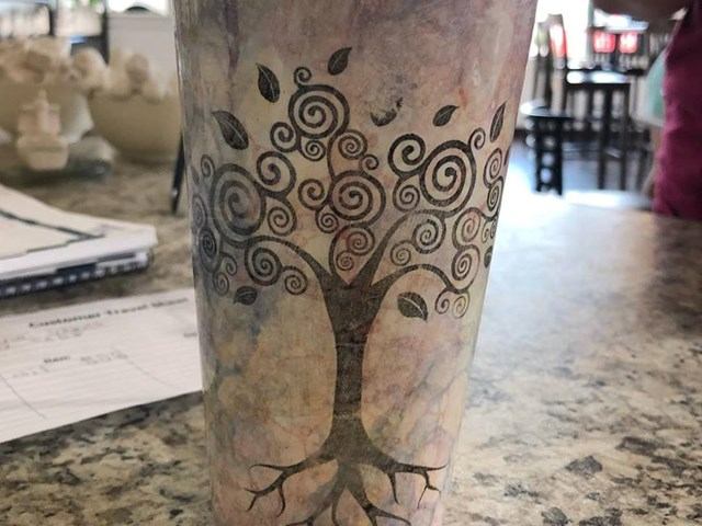 Painted tumbler with swirling tree design on tabletop beside papers.