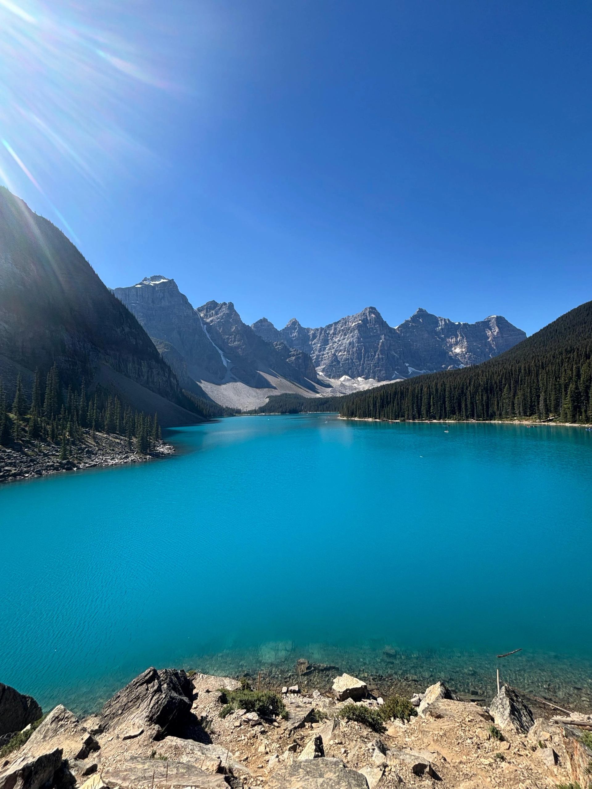 Turquoise lake with forested shore, rocky edges, and snow-capped mountains.