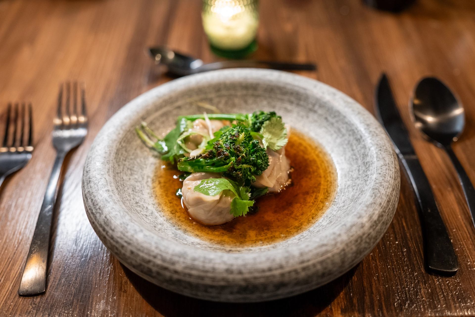 A dish of dumplings, broccoli, and herbs in broth, served in a gray stone bowl on a wooden table.
