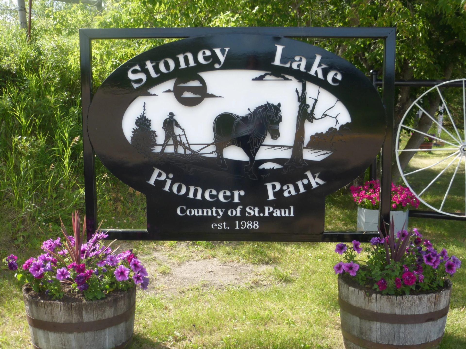 The image shows a park entrance sign surrounded by flower planters, trees, and greenery.