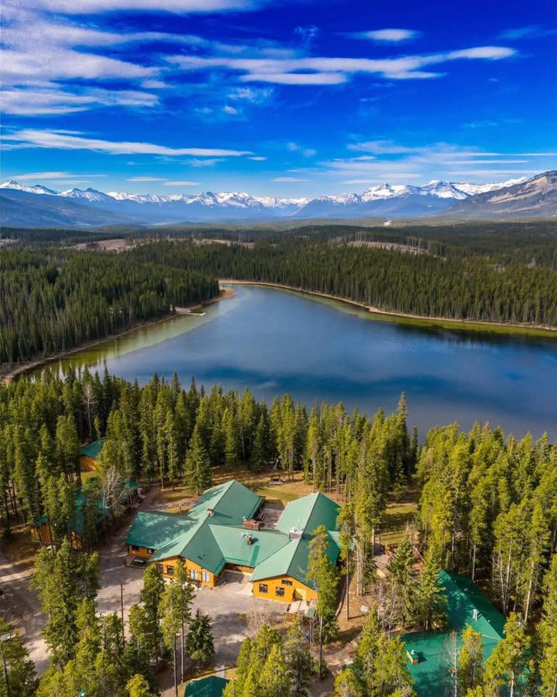Lakeside lodge with green roofs surrounded by dense forest and distant snow-capped mountains.