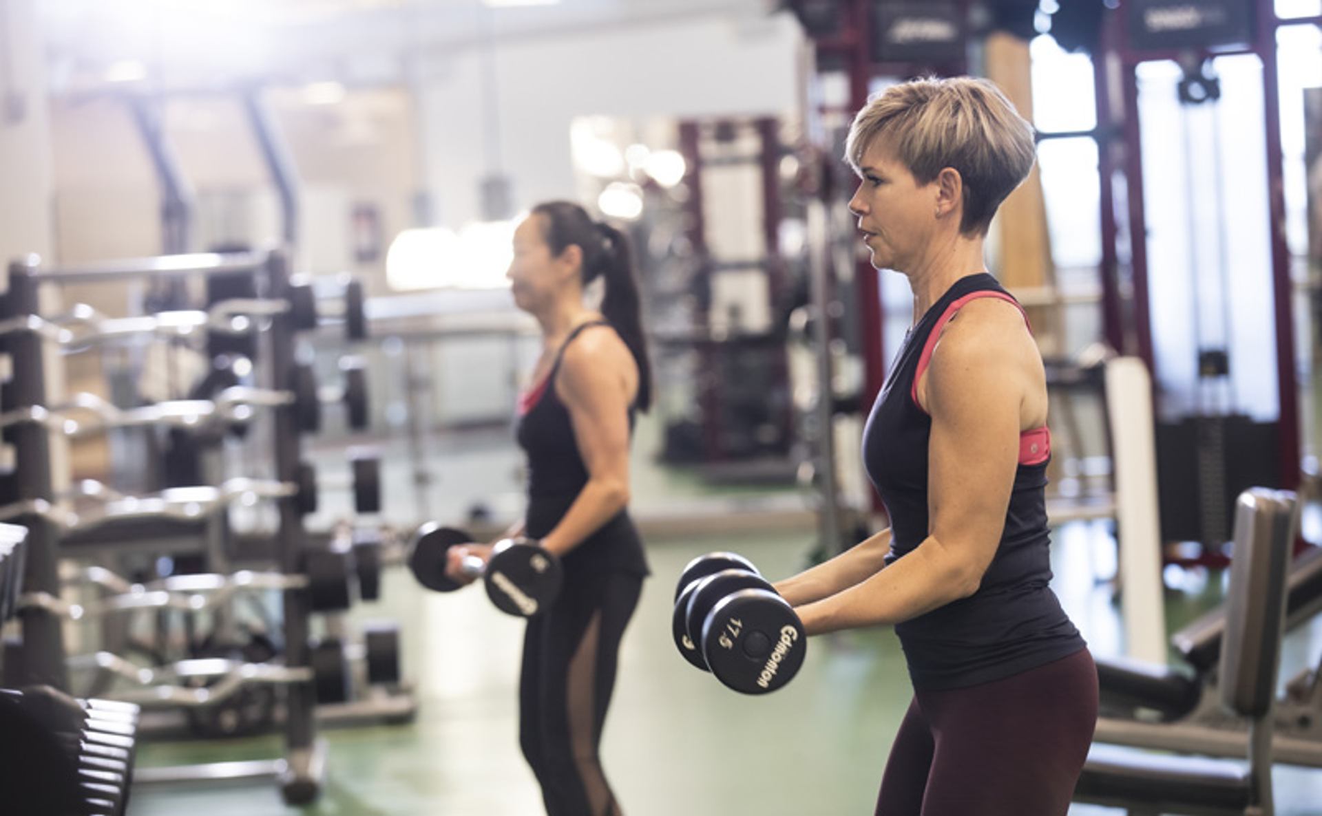 Two people lift dumbbells in a gym, surrounded by weight racks and machines.