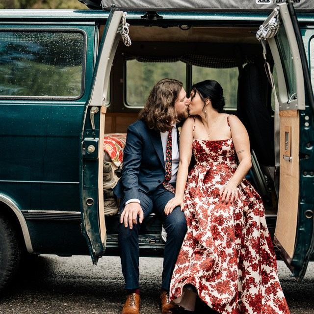 Wedding couple sits together in the open back of a vintage van, dressed in formal attire outdoors.