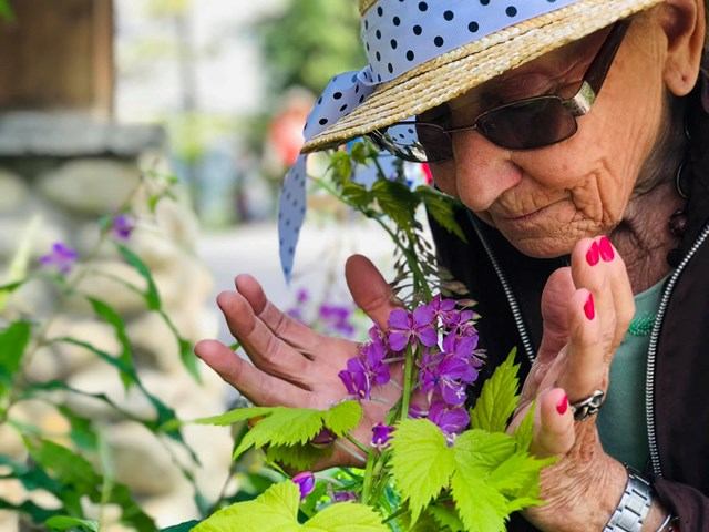 Person in polka-dot hat and sunglasses leaning toward purple flowers in a garden.