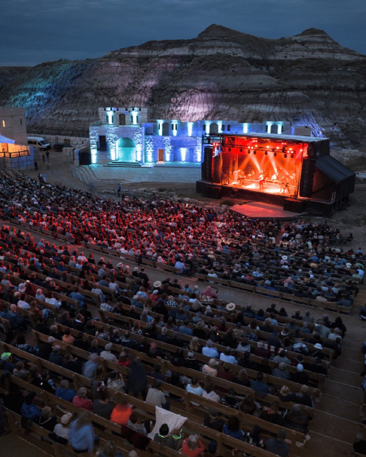 Outdoor amphitheatre crowd watching illuminated stage performance against rocky hillside backdrop.