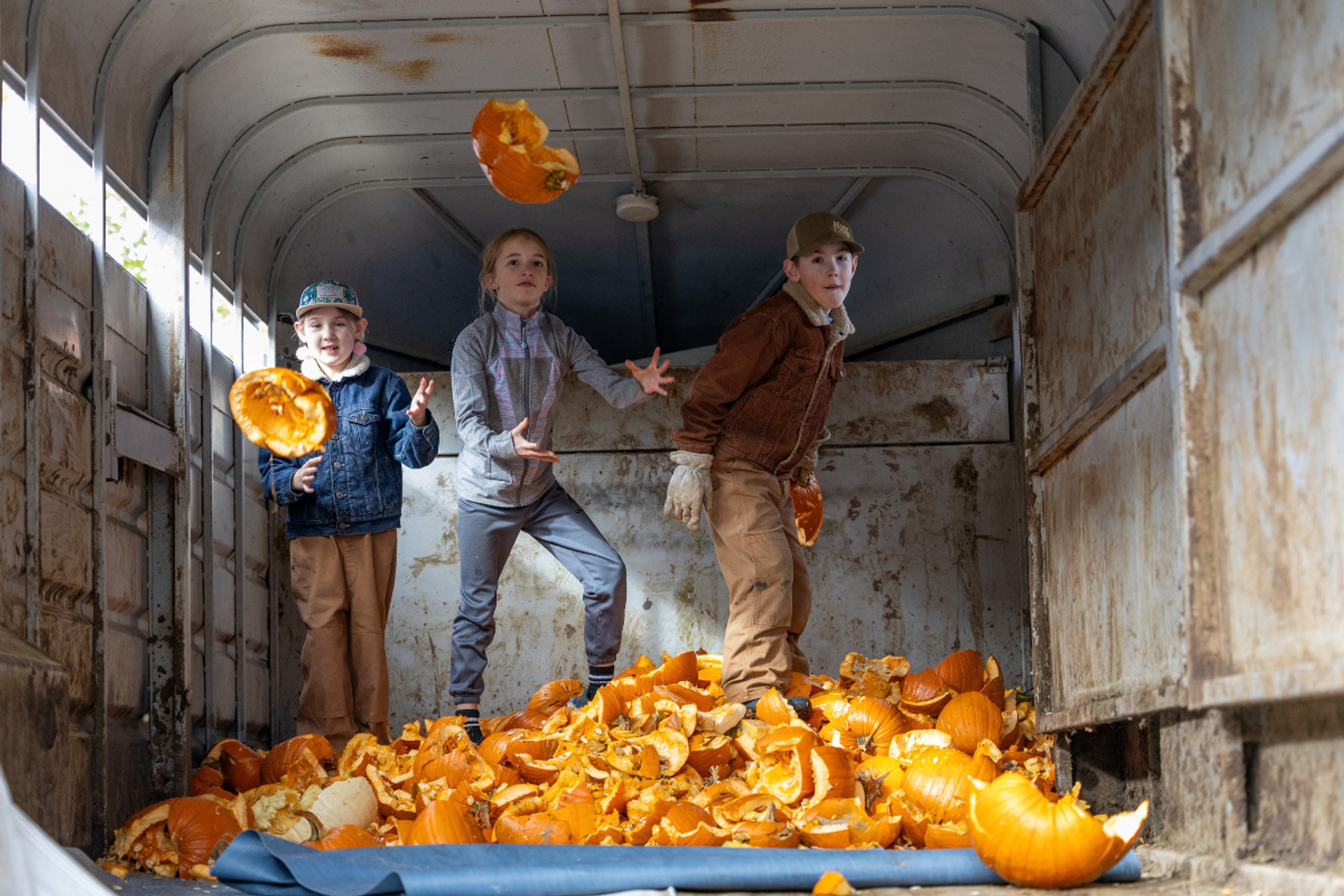 Three children toss broken pumpkins inside a weathered trailer.