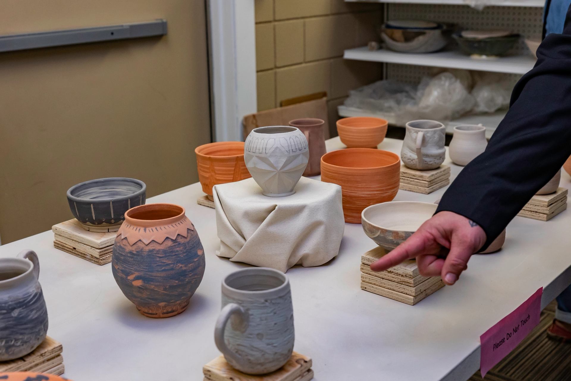 Ceramic mugs, bowls, and vases on display with hand pointing at one piece.