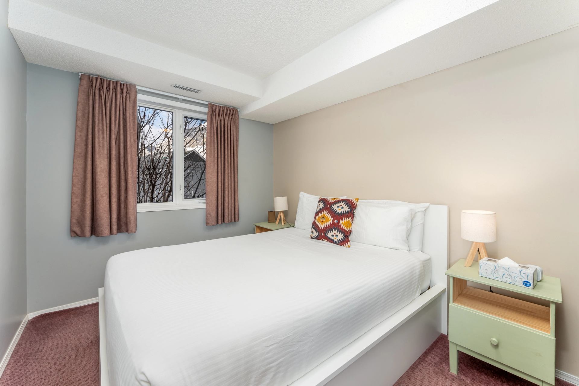 Small bedroom with white bed, patterned cushion, window, and two bedside tables.