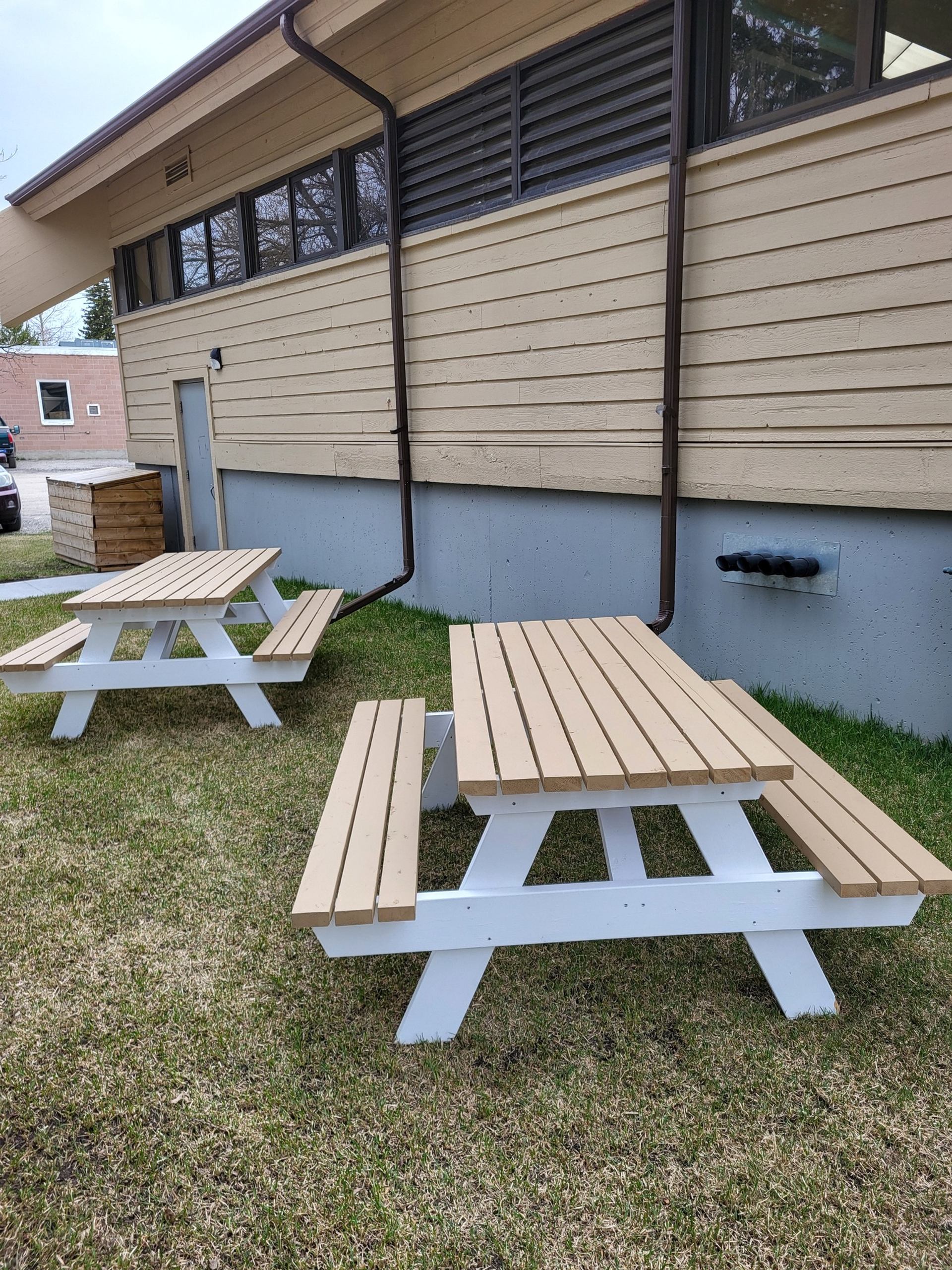Wooden picnic tables on grass beside beige building with high windows and downspouts.