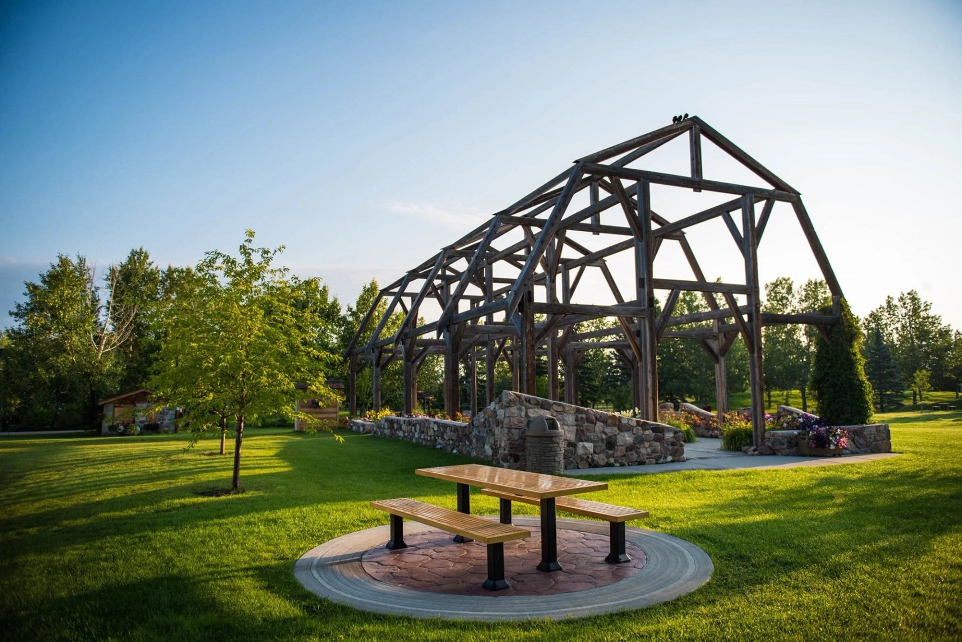 Wooden picnic table on grass near open timber pavilion and stone landscaping in park.
