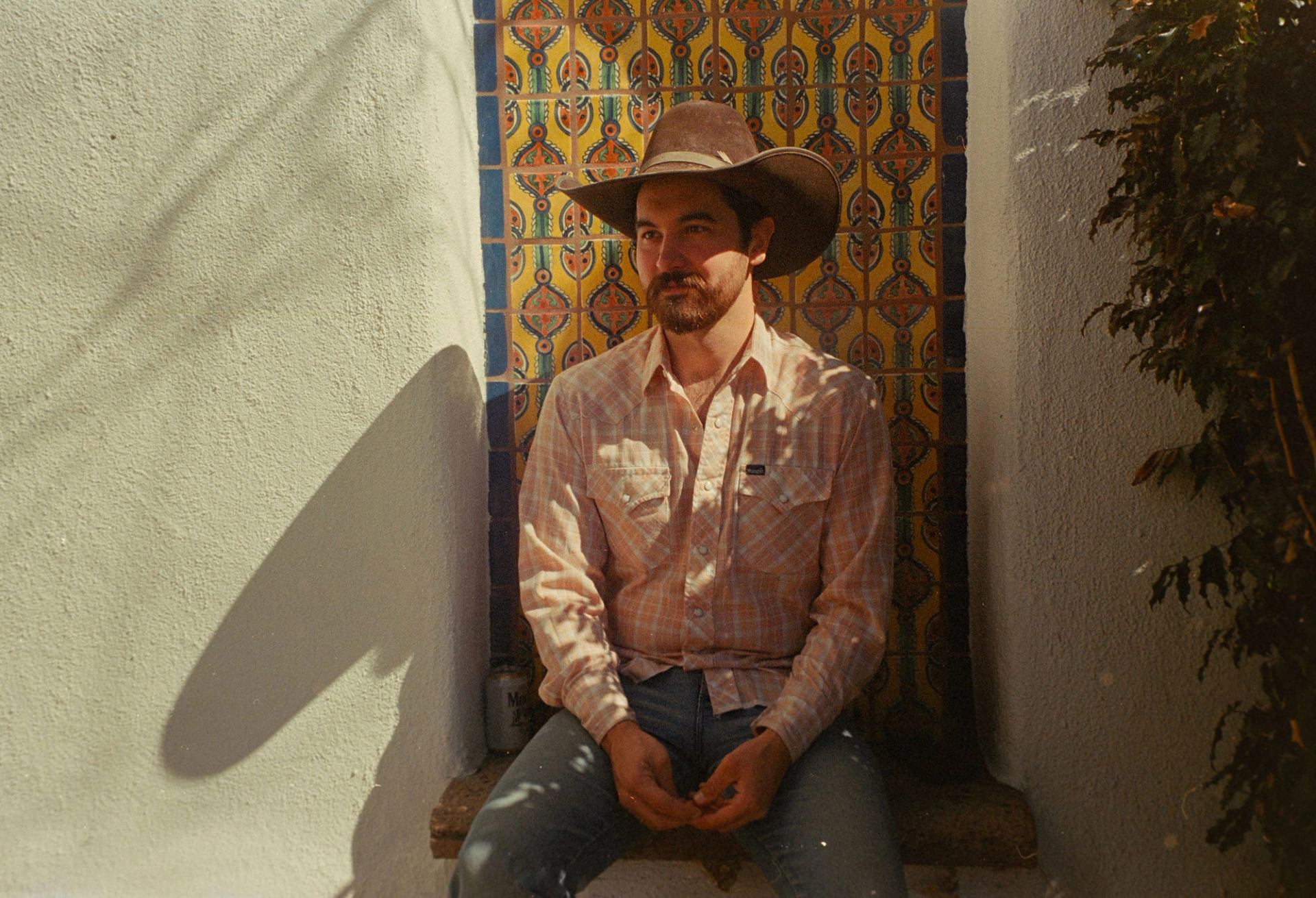 Person in cowboy hat sits against colourful tiled wall in narrow outdoor alcove.