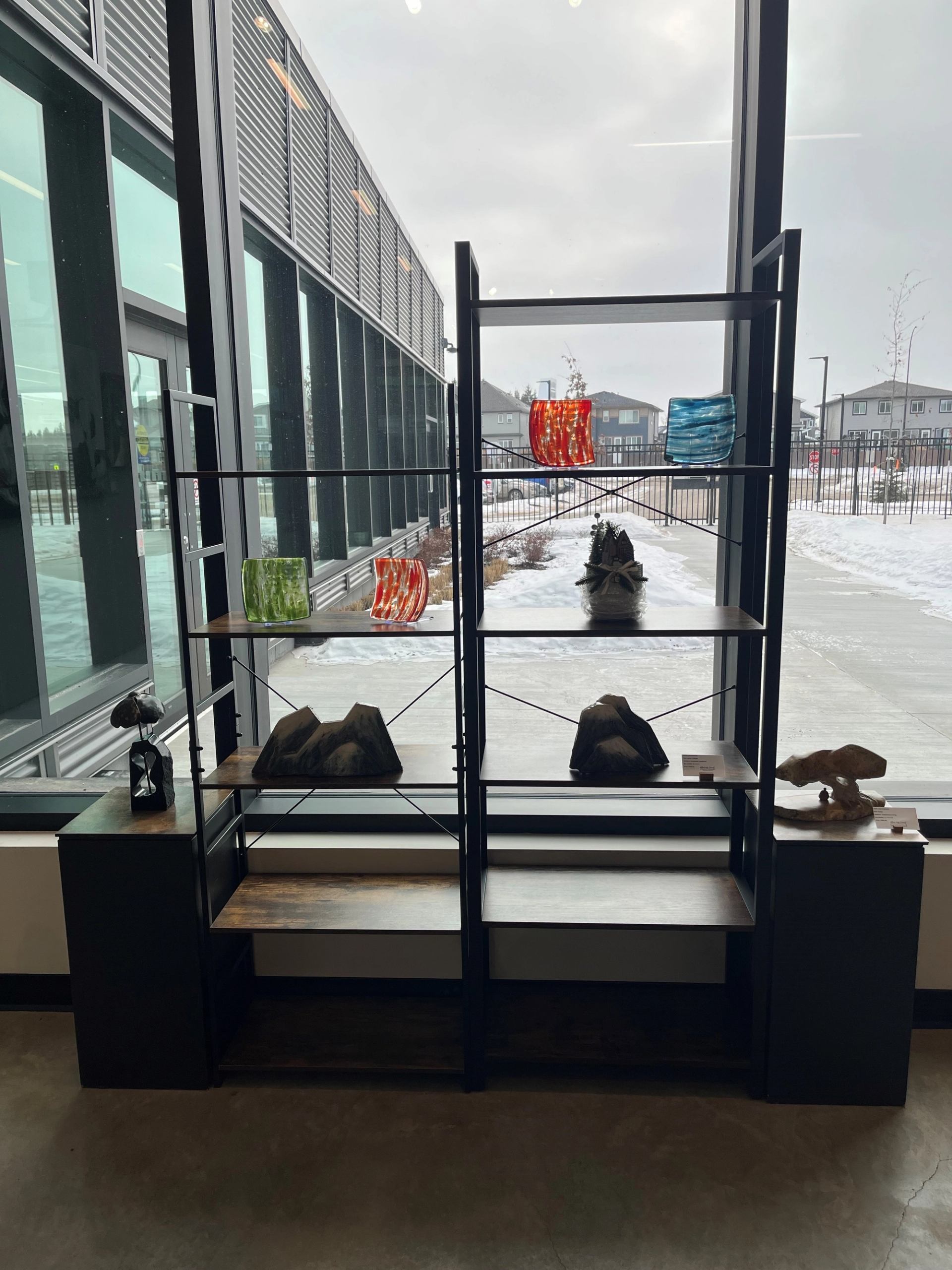Metal and wood shelving unit with glass art and sculptures beside large windows.