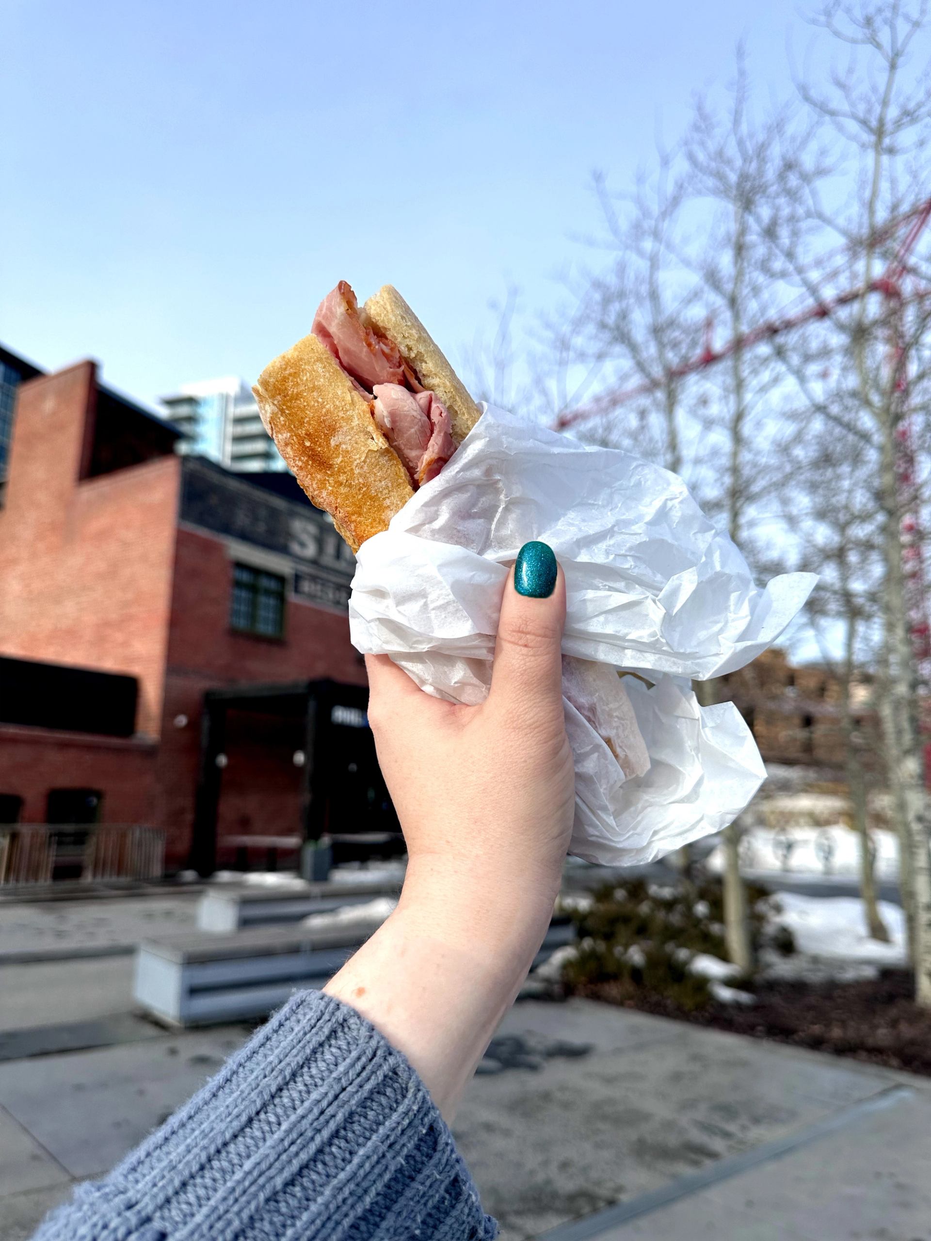Hand holding a bacon sandwich outdoors in a snowy urban setting.