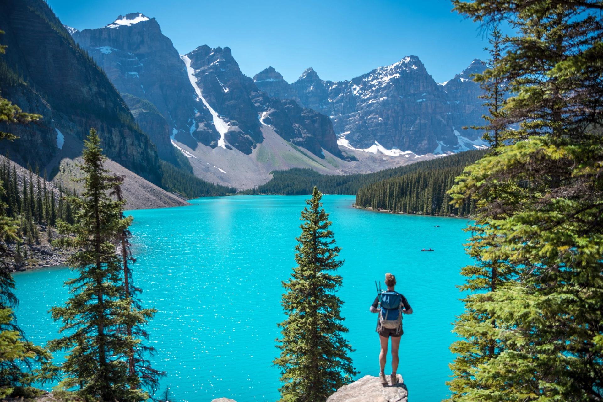 Person on rock overlooking turquoise lake, forest, and snow-capped mountains.