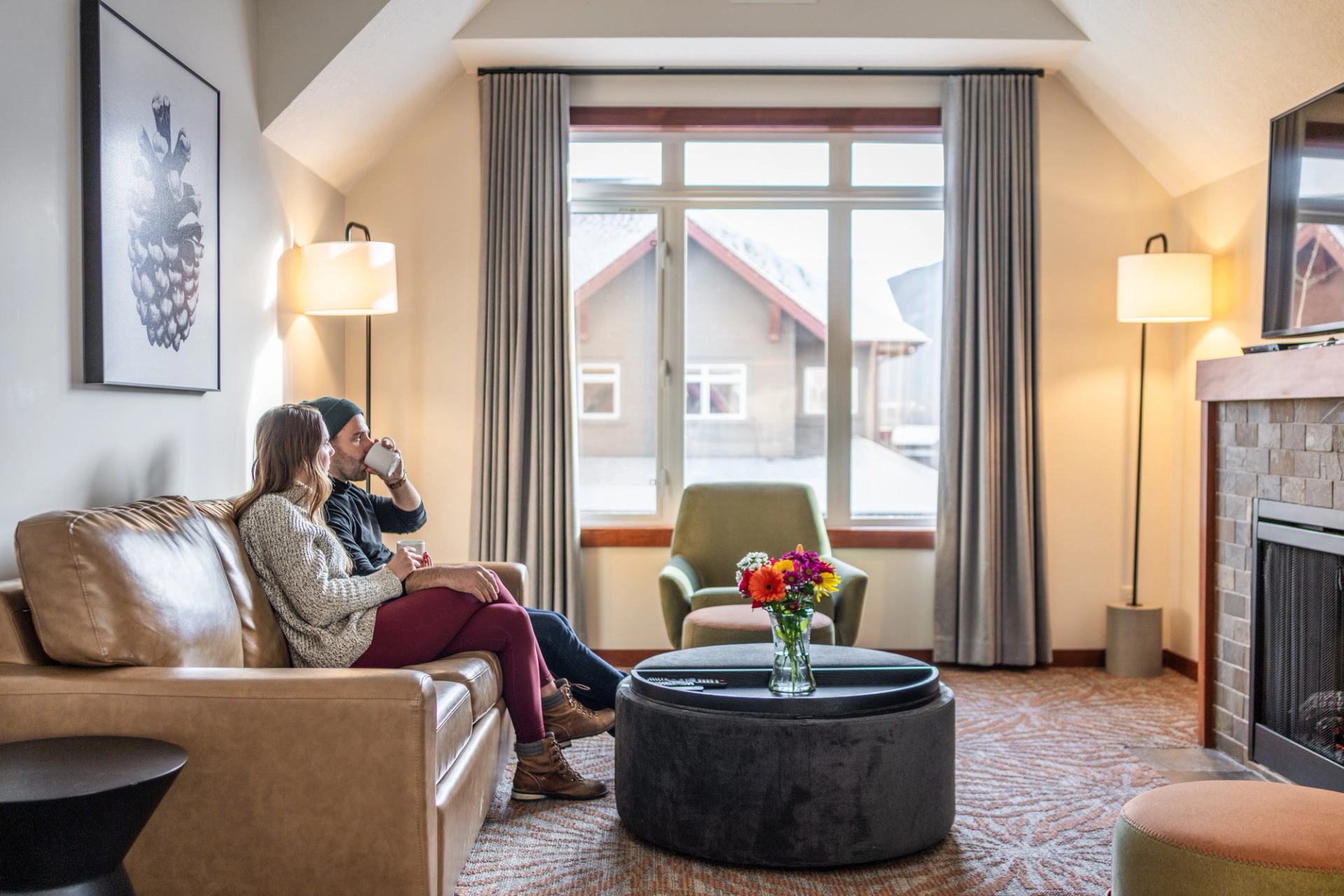 Two people sit on sofa with mugs beside fireplace and large window.