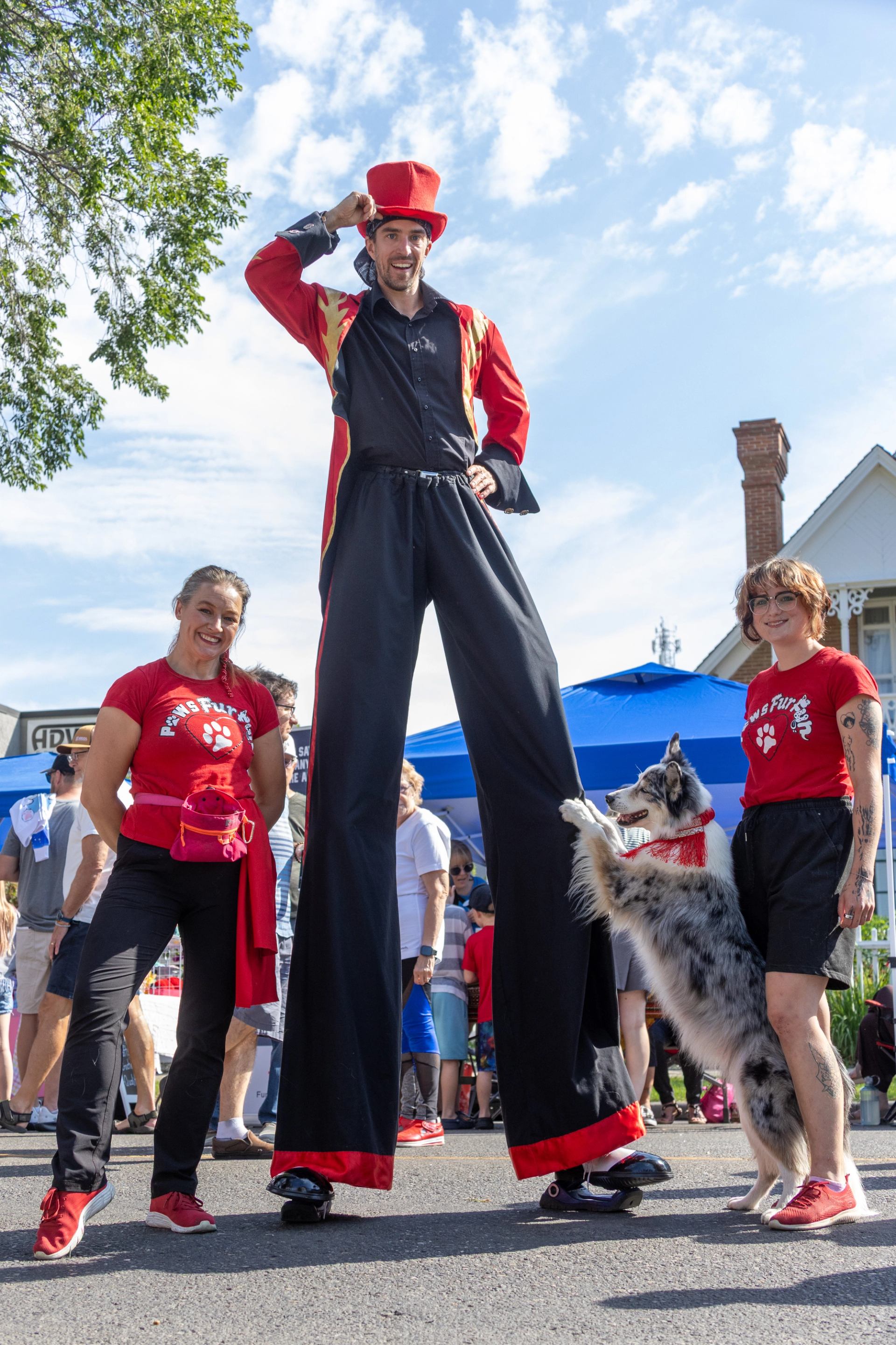Stilt walker in red costume stands between two people in red shirts and dog.